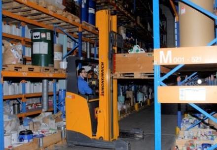Gabelstaplerfahrer im Lager zwischen hohen Regalen mit Kartons und Fässern
